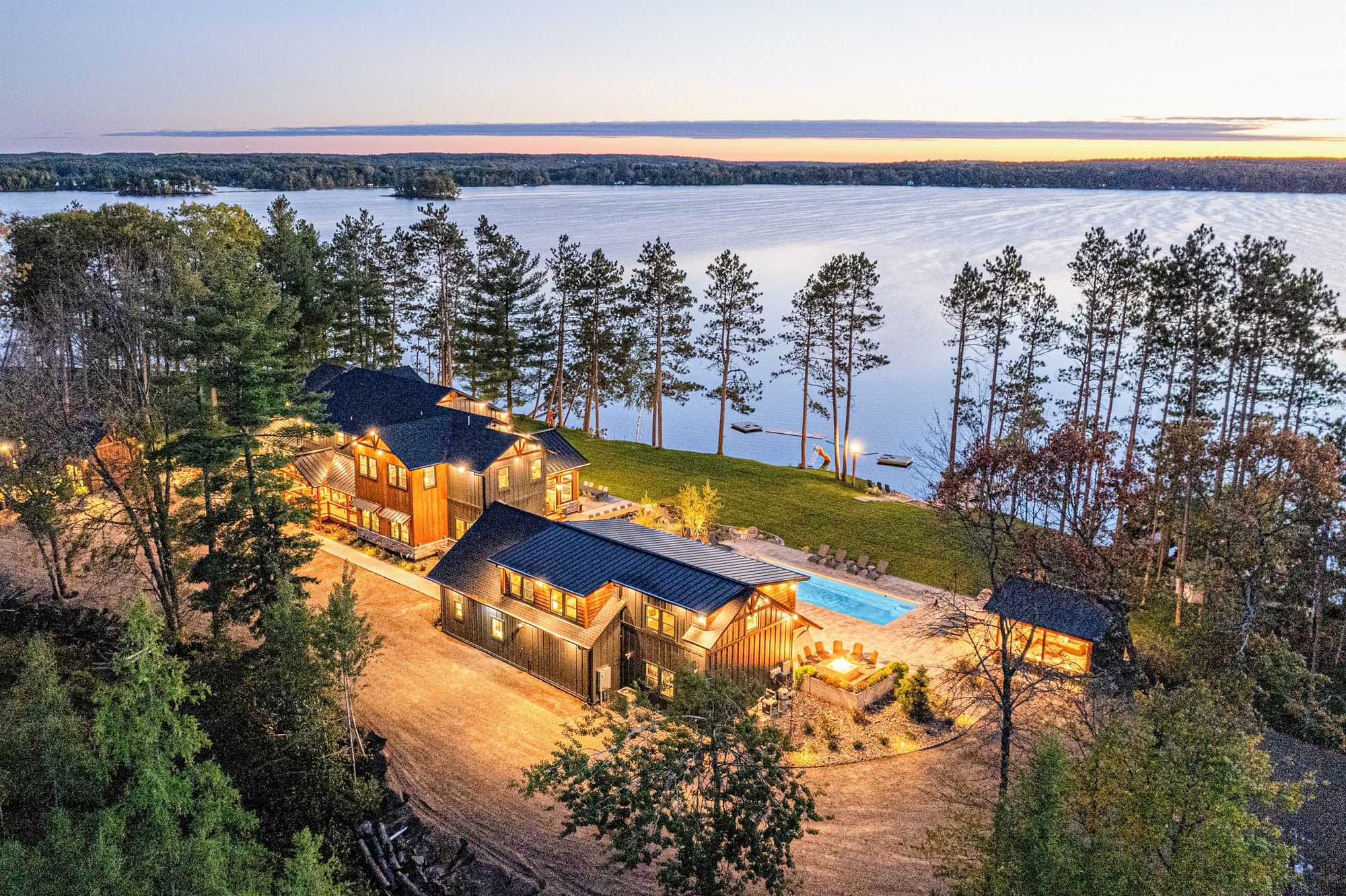 Aerial view of the Spooner Lake island lodge at dusk, lights glowing beside the pool and the lake beyond